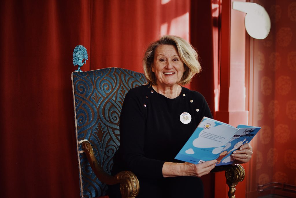 En leende sagotant i svart tröja sitter i en blå kungastol och läser ur en bok om Alfons Åberg. En glad sagotant sitter i en blå kungastol och läser ur en bok om Alfons Åberg.