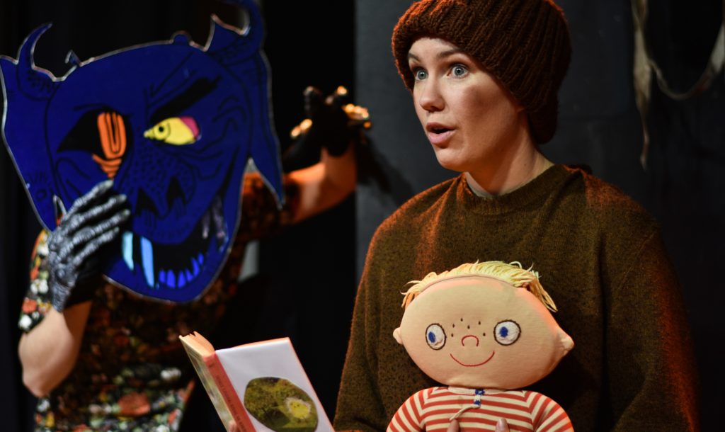 En person håller en docka och läser en saga. En mask föreställande monster syns vid sidan om.